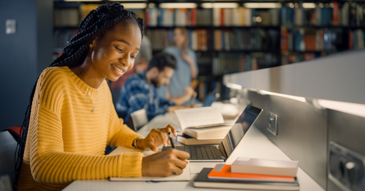 Student in a yellow sweater smiles while using a laptop and notebook at a library desk with books nearby.