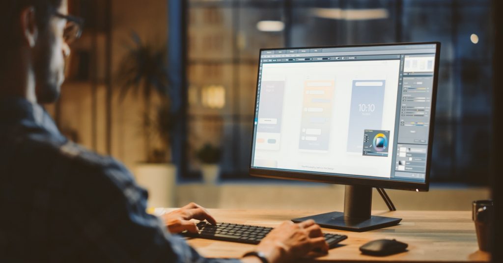 Designer works at a desktop monitor showing app layouts and color tools in a dimly lit office at night.