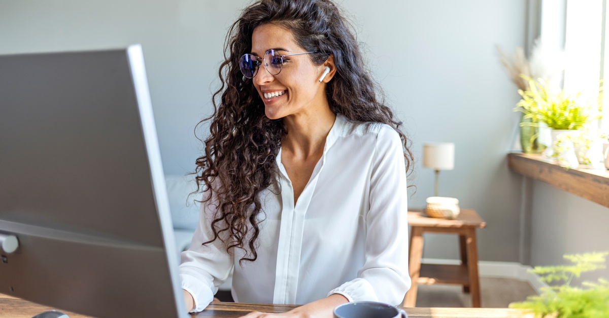 Woman with curly hair, glasses, and earbuds smiles while working at a desktop computer beside a sunny window.