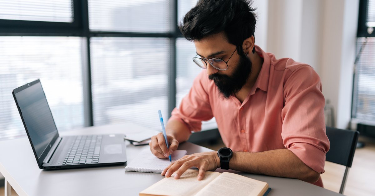 A man wearing glasses takes handwritten notes from an open book with a laptop open on the desk nearby.