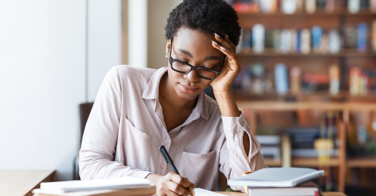 Woman wearing glasses takes handwritten notes in a notebook at a library table with books stacked on both sides.