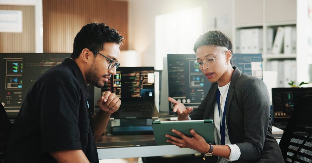 Two people discuss information on a tablet at a workstation with multiple screens showing code and software interfaces.