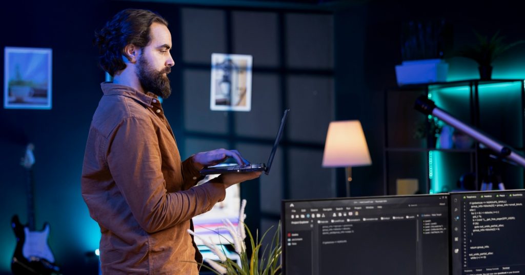 Man uses a laptop in a dark workspace with glowing blue lights, plants, and screens displaying code.