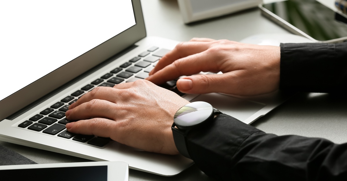 Hands type on a laptop beside a tablet, with a round smartwatch visible on the wrist at a desk.