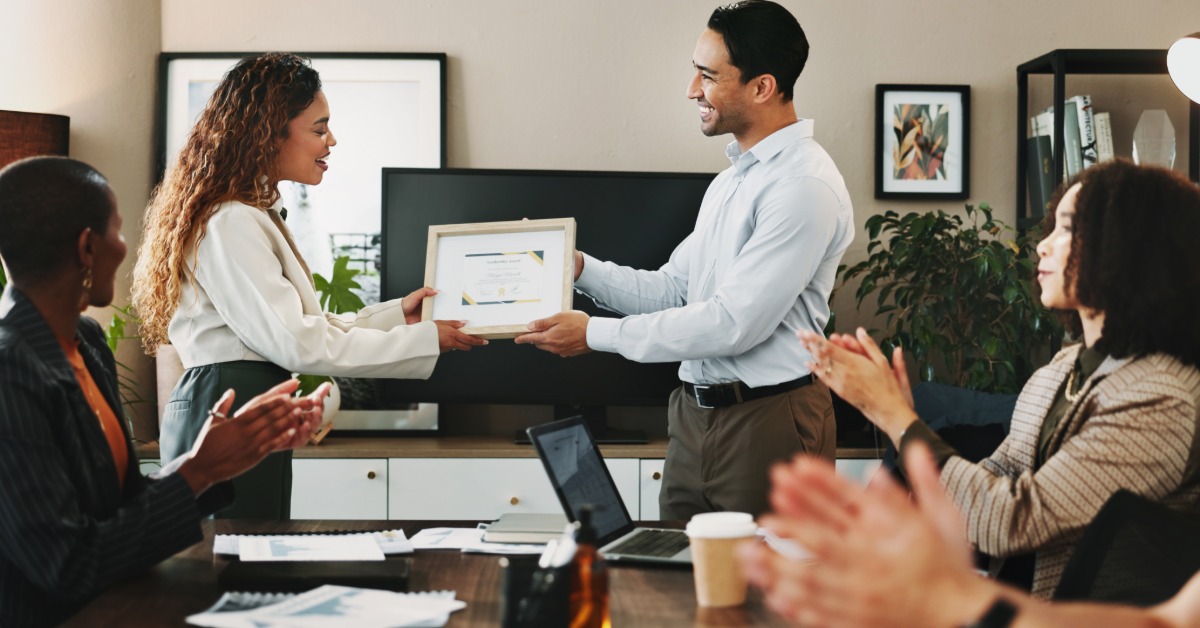Smiling coworkers exchange a framed award at the front of a conference room while teammates applaud.