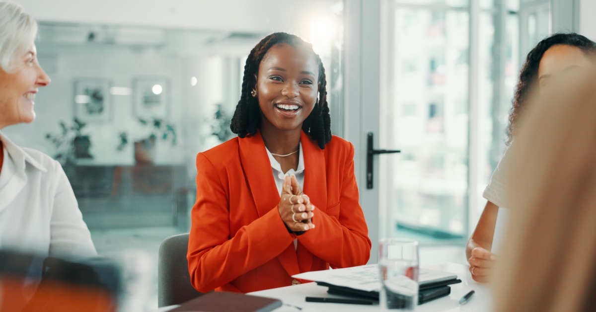 Smiling woman in a bright orange suit speaks with colleagues gathered around a table in a modern office.