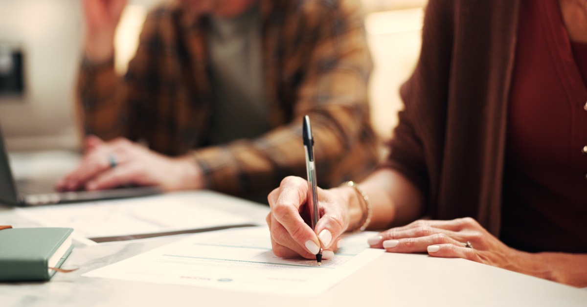 Hand writing on a printed document during an office meeting, with another participant blurred in the background.