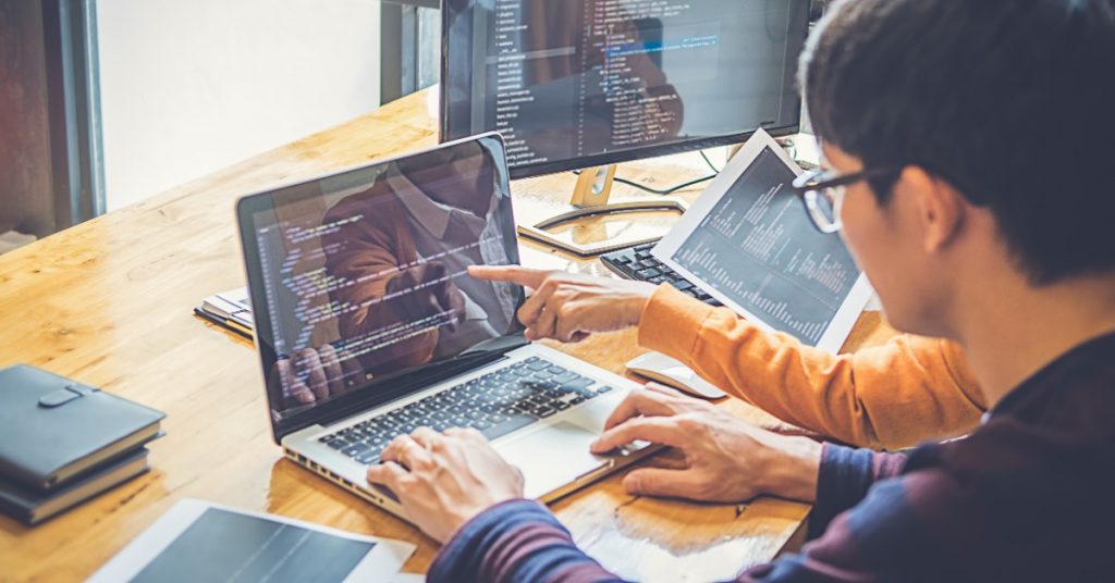 Developers review programming code together on multiple screens while working at a shared desk beside a large window.