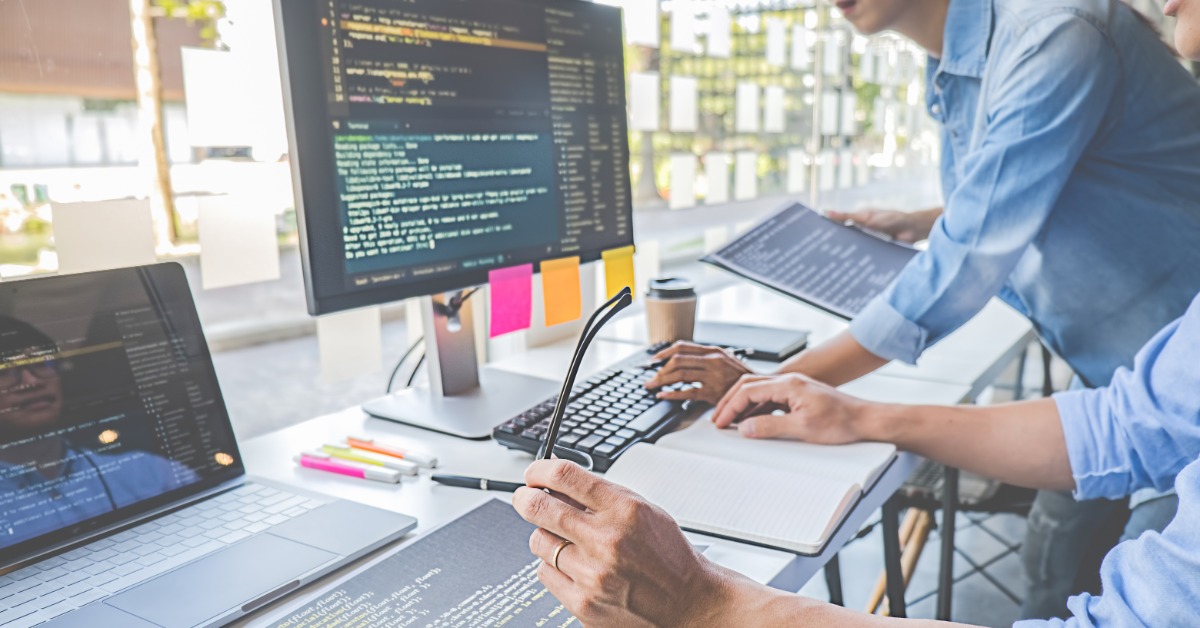 Two people work at a desk with computer screens displaying code, using a keyboard, laptop, and printed documents.