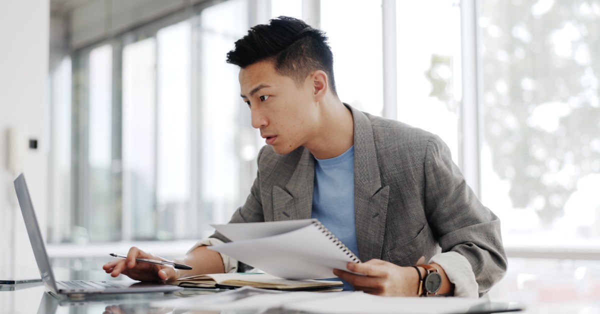 Man studies documents beside an open laptop, holding a pen and papers while seated at a bright office desk.