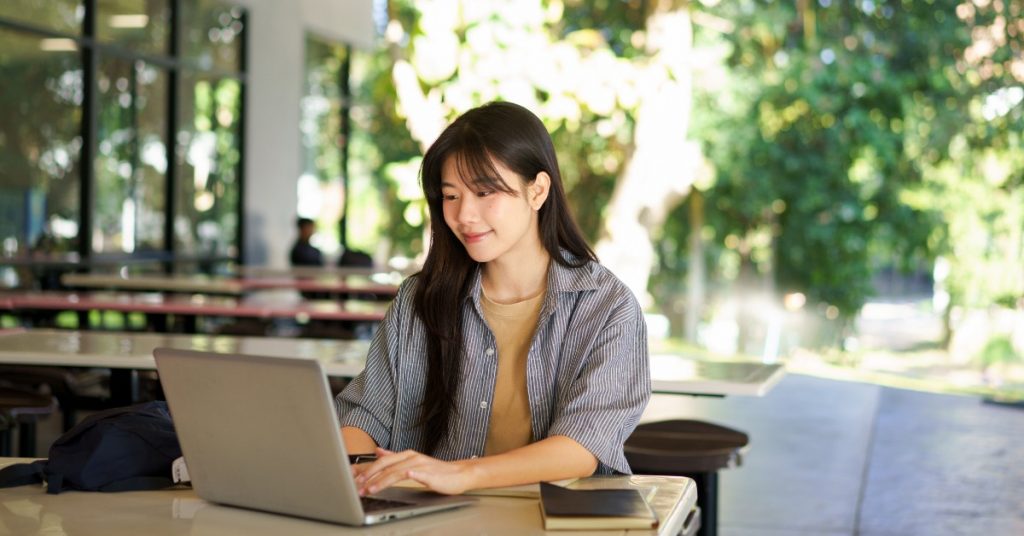 Woman types on a laptop at a covered outdoor study area, with a notebook on the table and green trees in the background.