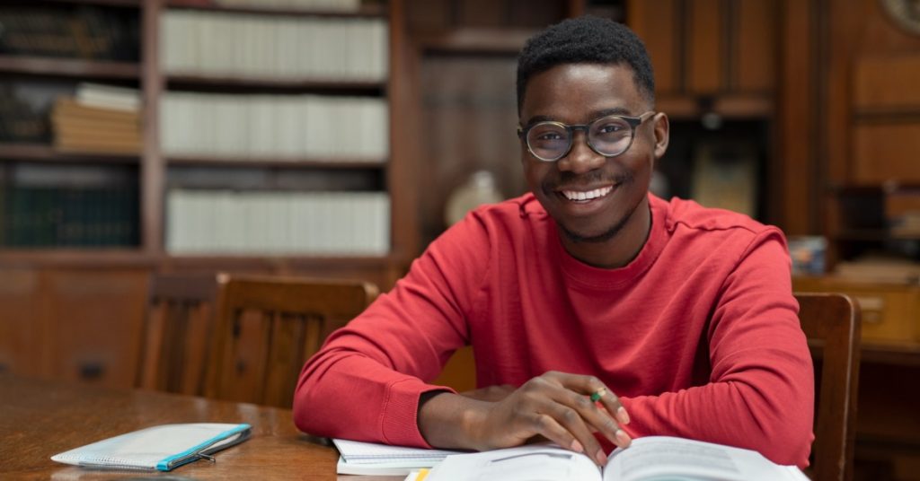 Man wearing glasses studies at a wooden library table, smiling with an open textbook and notebook in front of him.