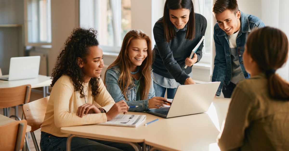 Group of students collaborate around an open laptop in a bright classroom, with several seated and others standing.
