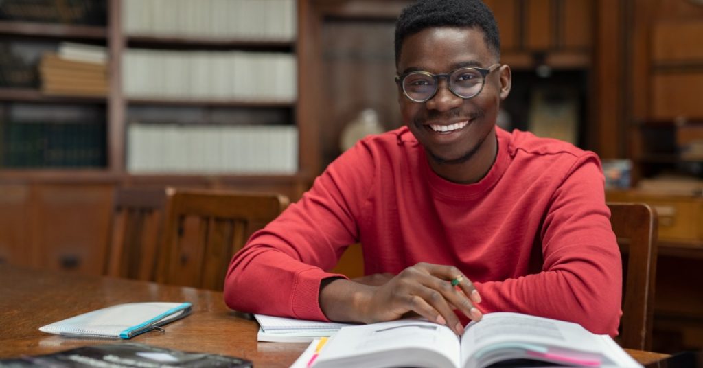 Student wearing glasses smiles at a library table while studying from an open textbook with notes and a pencil.