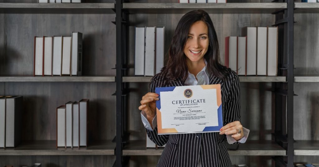 Woman smiles while displaying a certificate of appreciation in front of dark bookshelves filled with white books.